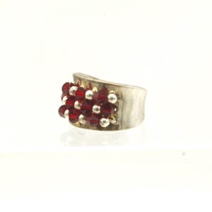 May include: A silver-toned ring with a cluster of small, deep red beads and silver-colored beads. The ring has a wide, curved band and a textured surface. The beads are arranged in a dense, textured pattern, creating a striking contrast.