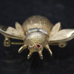 May include: A gold-toned bee brooch with red and white rhinestones for eyes. The bee has a textured body and wings.