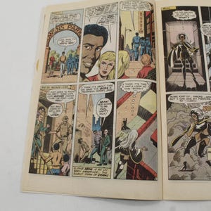 May include: Open comic book pages featuring colorful panels. The panels depict various scenes with characters in action, including a woman with white hair and a superhero costume. Text bubbles and captions are present.
