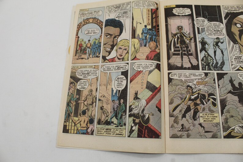 May include: Open comic book pages featuring colorful panels. The panels depict various scenes with characters in action, including a woman with white hair and a superhero costume. Text bubbles and captions are present.