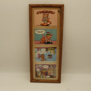 May include: A wooden picture frame with three Garfield comic strips. The top comic strip says "G'day Mate!" The middle comic strip says "Let's do lunch sometime." The bottom comic strip says "What's happening?"