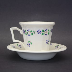 May include: White teacup and saucer set with a floral pattern of blue flowers and green leaves. The teacup has a square handle and a slightly flared rim. The saucer has a matching floral design around the edge.