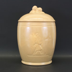 May include: A tan ceramic jar with a lid featuring a raised design of a person walking. The jar has a smooth, glossy finish.