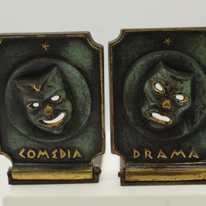 May include: Pair of antique bookends featuring comedy and drama masks. Each bookend has a dark green finish with gold accents. The comedy mask has a smiling face, while the drama mask has a frowning face. The word "COMEDIA" is on one bookend and "DRAMA" on the other.