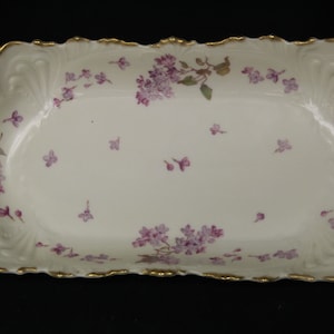 May include: A white porcelain serving dish with a scalloped edge and gold trim. The dish is decorated with clusters of purple lilac flowers and scattered individual blossoms. A decorative piece for serving food.