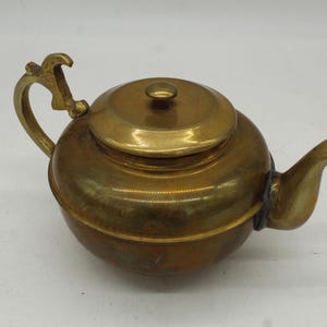 May include: A vintage brass teapot with a rounded body, a curved spout, and a decorative handle. The lid has a small knob. The teapot has a warm, golden color and appears to be made of metal. It is likely used for brewing tea.