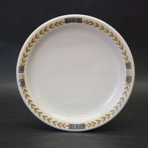 May include: A white ceramic plate with a gold and black border. The border features a repeating pattern of leaves and geometric shapes.