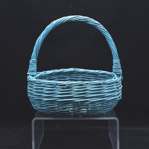 May include: A teal-colored woven basket featuring a rounded base and a single arched handle. The basket is constructed from thin, flexible material woven in a horizontal pattern, with the handle securely attached to each side of the rim.