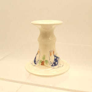 May include: A cream-colored ceramic candle holder with a wide base and a flared top. The holder is decorated with two white rabbits wearing blue and white patterned bibs, with a carrot in between them. The design is hand-painted.