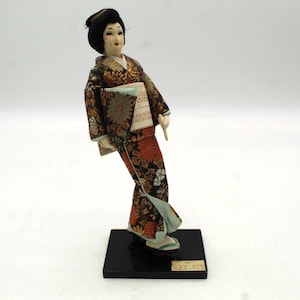 May include: A Japanese doll in a traditional kimono, standing on a black base. The kimono features a floral pattern in shades of red, gold, and black. The doll has a serene expression and is a collectible item.
