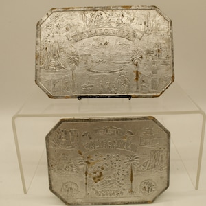 May include: Two silver-toned metal plaques with an embossed design of California landmarks. The top plaque reads "CALIFORNIA" and the bottom plaque reads "CALIFORNIA".
