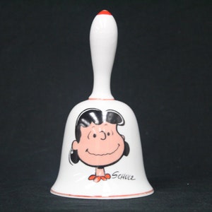 May include: White ceramic bell with a red handle and a black and white illustration of a cartoon girl with black hair and red shoes. The bell has the signature "Schulz" on the bottom.