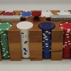 May include: A wooden poker chip set with a variety of colored chips, including red, green, blue, and white. The chips are stacked in columns within a wooden holder. A red chip is on top of the holder.