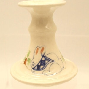 May include: A white ceramic candlestick holder with a hand-painted design of a rabbit wearing a blue and white checkered scarf. The rabbit is next to a small green and orange plant.