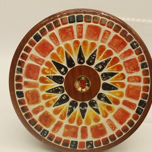 May include: A round, mosaic-tiled decorative object with a wooden frame. The mosaic features a sunburst pattern in shades of red, orange, yellow, and black. The center has a small, round, wooden accent with a red gem.