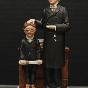 May include: A porcelain figurine of a man in a black suit and a boy in a black suit holding a scroll. The man is pointing at the boy and the boy is looking at the scroll. The figurine is on a white base with the words "Norman Rockwell" written on it.