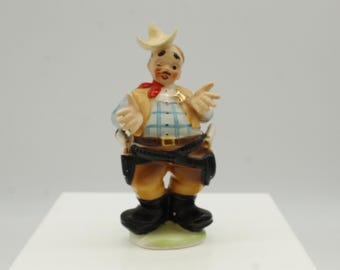May include: A ceramic figurine of a cowboy with a cream-coloured hat, a red neckerchief, and a gold star badge. The cowboy wears a plaid shirt, tan vest, and brown trousers. Black boots and holsters complete the outfit.