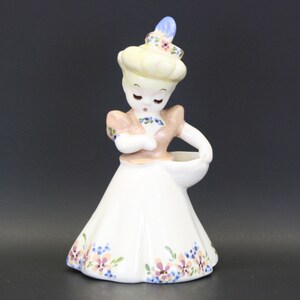 May include: A ceramic figurine of a girl wearing a pink dress and a floral hat. She is holding a small white planter in her hand. The figurine is decorated with floral details in shades of blue, pink, and green.