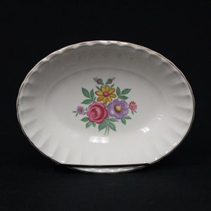 May include: White oval serving dish with a floral bouquet design. The bouquet features pink, yellow, and purple flowers with green leaves. The rim has a scalloped edge and a gold trim. The dish is displayed on a black stand.
