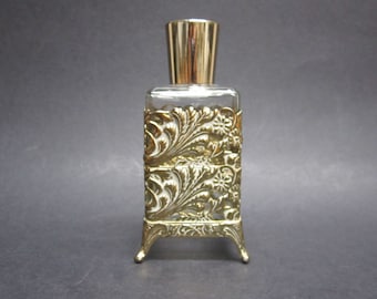 May include: A rectangular glass perfume bottle featuring a gold-toned metal filigree overlay with intricate floral and leaf patterns. The bottle stands on four small metal feet and is topped with a smooth, cylindrical gold-toned metal cap.