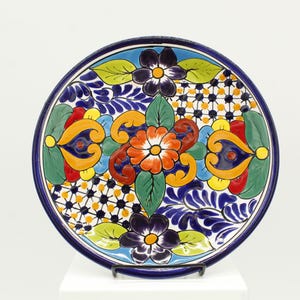 May include: A hand-painted ceramic plate with a vibrant floral design. The plate features a dark blue rim and a central pattern of orange, red, yellow, and blue flowers and leaves. The plate is approximately 10 inches in diameter.