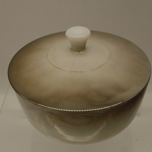 May include: A small, round, silver-colored container with a white lid and knob. The container has a smooth, shiny surface.