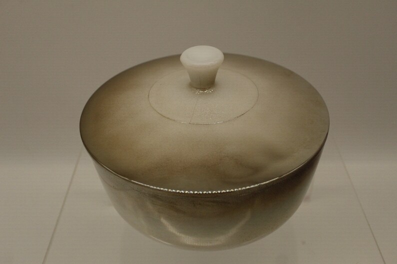 May include: A small, round, silver-colored container with a white lid and knob. The container has a smooth, shiny surface.