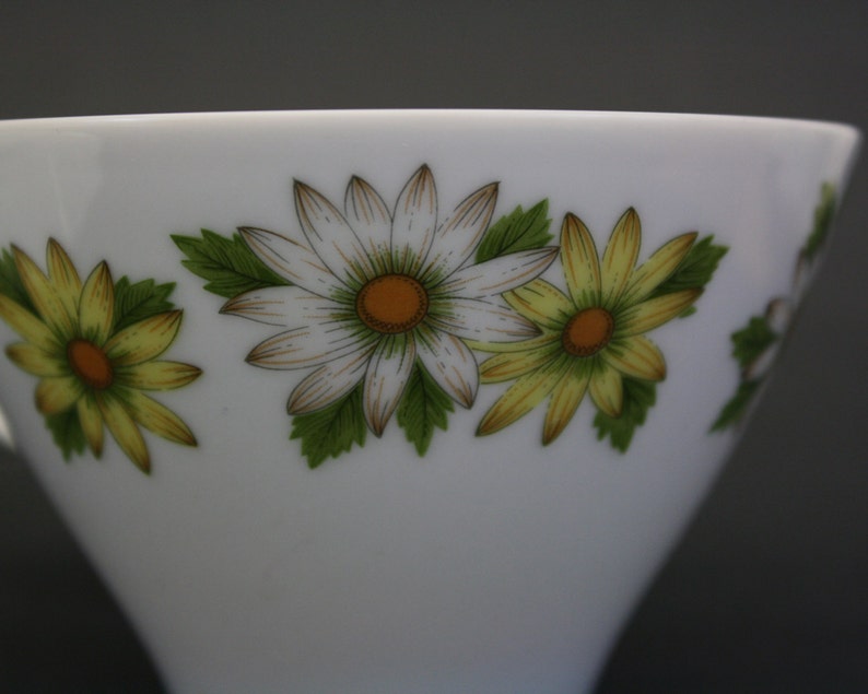 Puede incluir: Taza de t&eacute; blanca con un dise&ntilde;o floral de margaritas. Las margaritas tienen centros amarillos, blancos y naranjas con hojas verdes. La taza es de porcelana y tiene una forma simple y elegante.