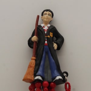 May include: A figurine of a young wizard wearing glasses, a black robe, and blue pants, holding a broom. The figure has a red and white striped shirt underneath the robe. A gold emblem is on the robe.