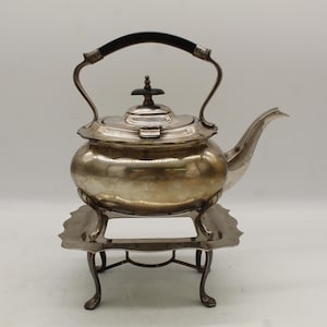 May include: A silver-colored teapot with a black handle and a decorative lid, resting on a matching stand. The teapot has a curved spout and sits on a decorative, ornate stand with four legs. The teapot and stand appear to be made of metal.