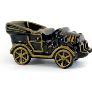 May include: Black ceramic vintage car shaped ashtray with gold accents. The car has a detailed design and is a great addition to any home decor.