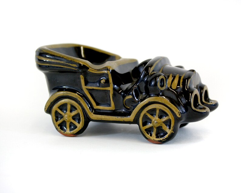May include: Black ceramic vintage car shaped ashtray with gold accents. The car has a detailed design and is a great addition to any home decor.