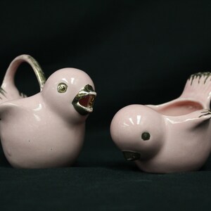 May include: Two pink ceramic bird figurines with gold accents. The bird on the left has its beak open and is holding a handle. The bird on the right is facing the left and has its beak closed.