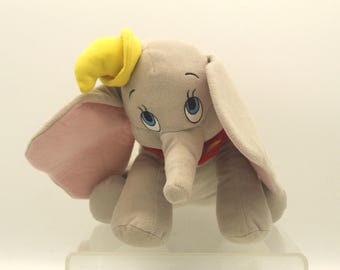 May include: A plush Dumbo the elephant toy. The elephant is gray with large, pink-lined ears, blue eyes, and a yellow hat. It has a red collar and is sitting down.
