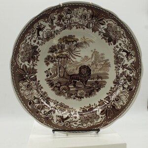 May include: A round ceramic plate featuring a brown and white transferware pattern. The center depicts a lion standing in a landscape with trees and mountains. The wide rim is decorated with a detailed floral border of various blossoms and leaves.
