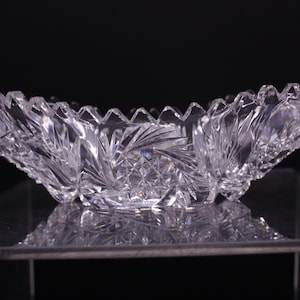 May include: A clear, cut-glass bowl with a scalloped rim. The bowl has a symmetrical, curved shape and intricate starburst and leaf-like patterns. The bowl is displayed on a reflective surface against a black background.