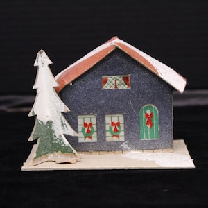 May include: A small blue paper house with a red roof and white trim. The house has three windows with red bows and a green door with a red bow. A small white and green paper Christmas tree is in front of the house.