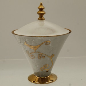 May include: A white ceramic sugar bowl with a gold lid and a gold bird design. The bowl has a conical shape and a gold base.
