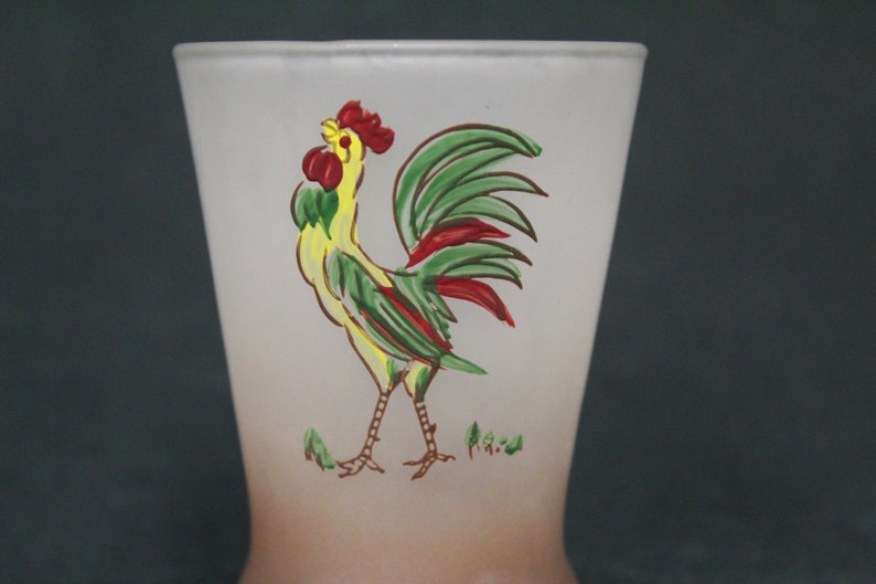 May include: A frosted glass drinking glass with a rooster design. The rooster is yellow, green, and red with a brown outline. The glass has a brown ombre effect at the bottom.