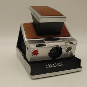 May include: A vintage Polaroid SX-70 Land Camera, featuring a silver body with brown leather accents. The camera has a distinctive folding design, a red button, and the "POLAROID SX-70 LAND CAMERA" logo on the front. It is a classic instant film camera.