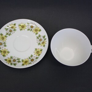 Puede incluir: Taza de t&eacute; blanca con asa y platillo a juego. El platillo presenta un patr&oacute;n floral de margaritas amarillas y blancas. La taza y el platillo est&aacute;n sobre una superficie oscura.