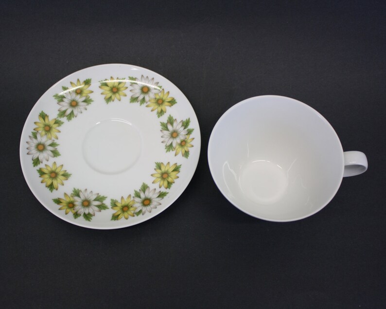 Puede incluir: Taza de t&eacute; blanca con asa y platillo a juego. El platillo presenta un patr&oacute;n floral de margaritas amarillas y blancas. La taza y el platillo est&aacute;n sobre una superficie oscura.