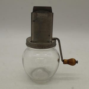 May include: A vintage, clear glass jar with a metal lid and a hand-cranked grater. The grater is made of metal and has a wooden handle. The jar is empty and sits on a white surface.