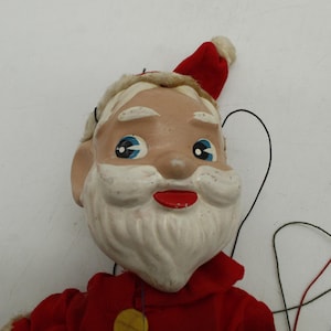May include: A vintage Santa Claus puppet with a red suit, hat, and a white beard. The puppet has blue eyes and a red mouth. Black strings and wooden control are visible. A classic Christmas decoration.