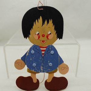 May include: Wooden pull-string toy in the shape of a boy. The toy has a painted face with rosy cheeks, a red nose, and a smiling mouth. He wears a blue jacket, a red and white striped shirt, yellow shorts, and brown shoes. The toy is suspended by a string.