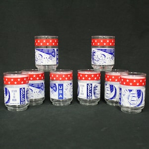 May include: A set of ten clear glass tumblers. Each glass features a red band with white stars at the top and a white and blue graphic with text related to the Apollo 12 mission. The glasses have a USA label.