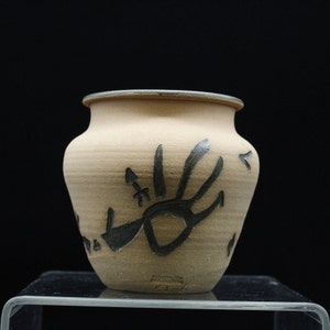 May include: A small, tan ceramic vase with a black handprint design. The vase has a rounded body and a slightly flared lip.