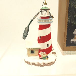 May include: A Christmas ornament shaped like a lighthouse with red and white stripes. The lighthouse has a small house attached, and figures of Santa Claus, a woman, and a penguin are at the base. The ornament has a green and white roof.