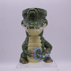 May include: A green ceramic alligator mug with a blue tail. The mug has a wide mouth and a textured surface.