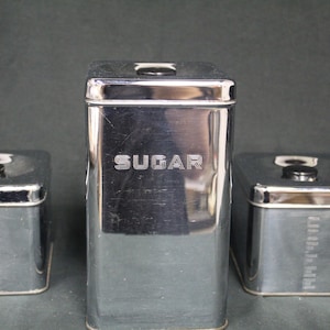 May include: A set of three vintage metal kitchen canisters. The tallest canister is labeled "SUGAR" in embossed lettering. The other two canisters are labeled "TEA" and "FLOUR". Each canister has a black knob on the lid.